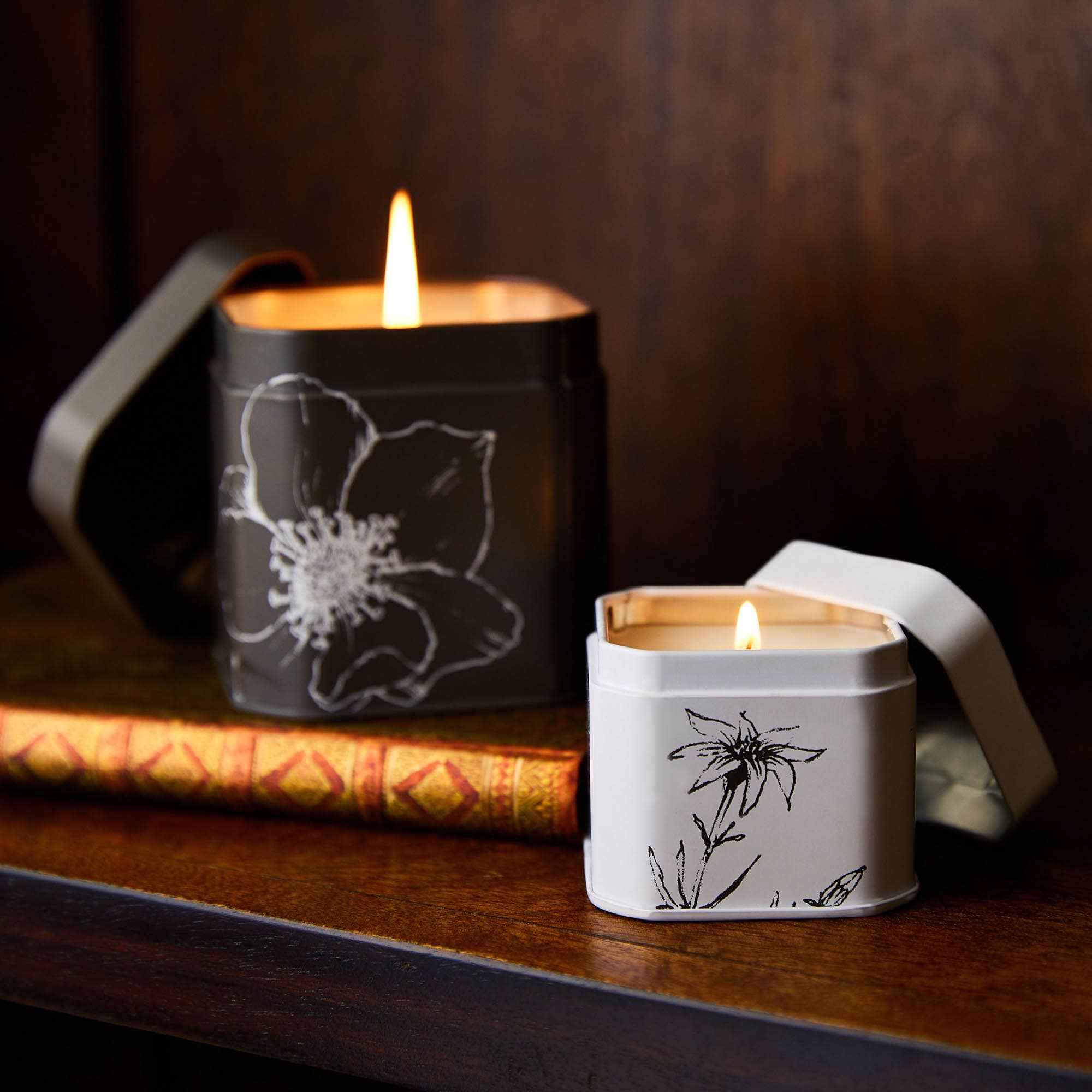 A white ceramic candle holder with black floral illustrations, lit with a small flame, placed on a wooden surface, creating a cozy, ambient atmosphere.