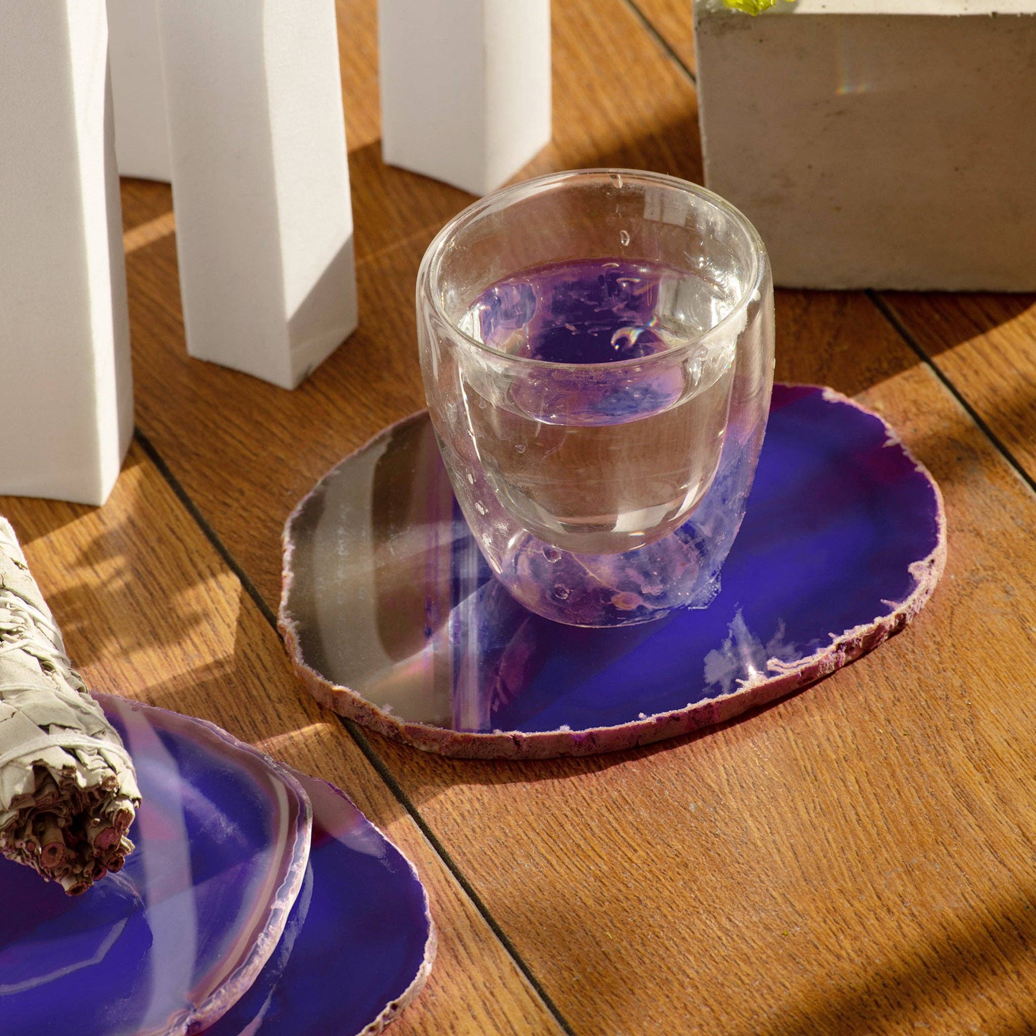 A glass of water with purple hues on a wooden table, placed on a purple agate coaster, surrounded by white decorative objects in theUnique Curiosities Collection for Home and Decor.