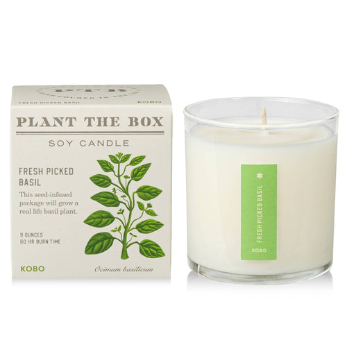 White soy candle with green basil plant illustration on the label, placed next to a basil plant and a matching plant-themed box, on a wooden surface.
