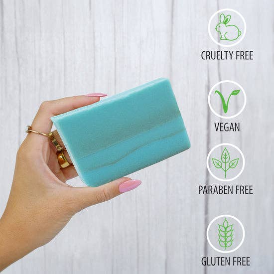 Hand holding a rectangular blue soap bar with icons indicating cruelty-free, vegan, paraben-free, and gluten-free features