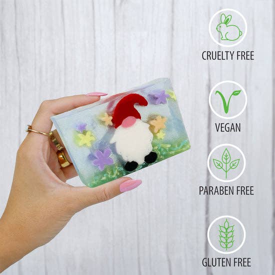 Hand holding a transparent soap package with garden gnome soap inside, surrounded by icons indicating cruelty-free, vegan, paraben-free, gluten-free