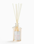 Elegant glass reed diffuser bottle with cream-colored reeds, tied with a satin ribbon, labeled with pink peony scent for home fragrance