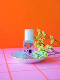 Nail polish bottle with playful astronaut and space-themed stickers, white cap, on a pink grid surface with yellow flowers and orange background