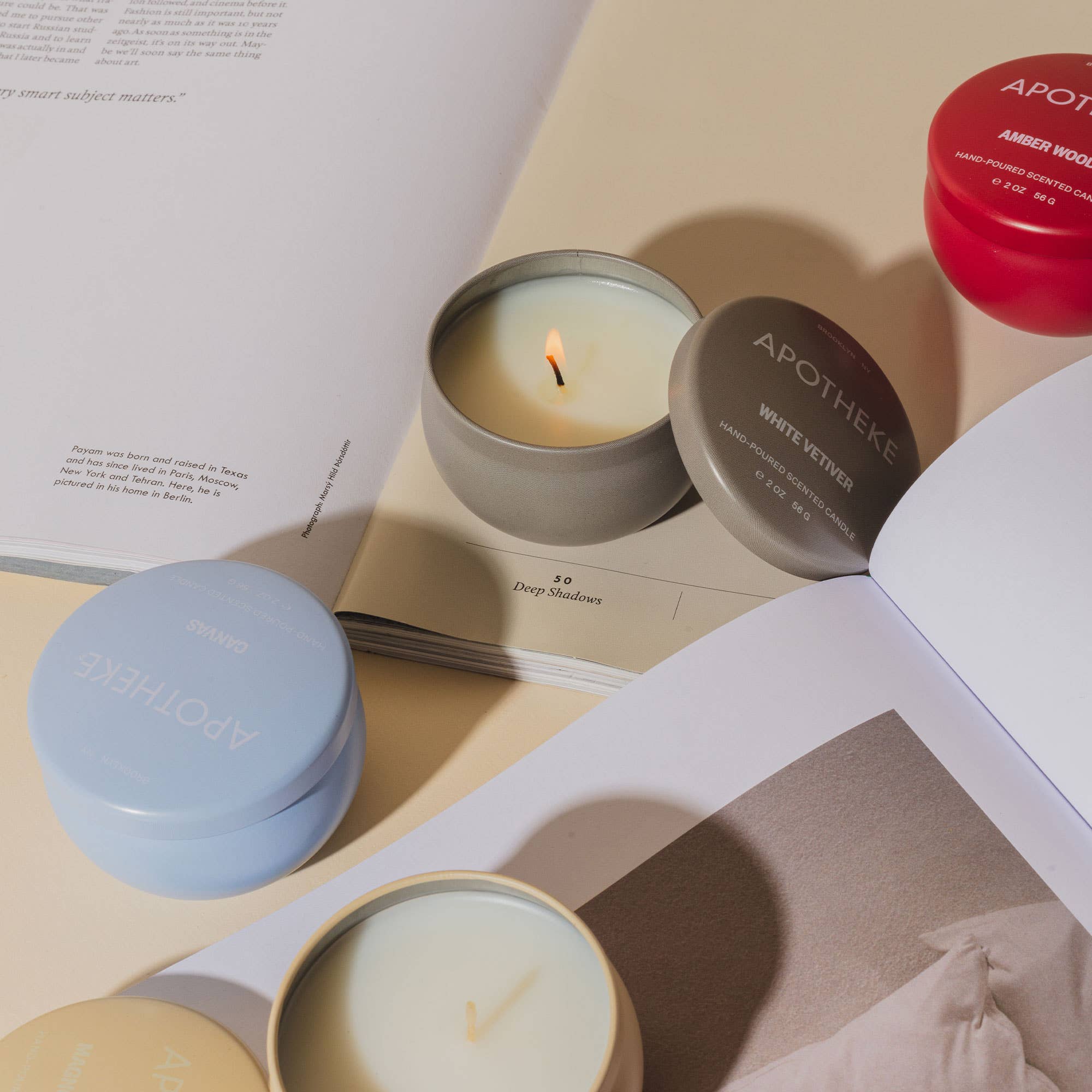 Open white candle with a lit wick, surrounded by various candle containers and an open book, creating a cozy ambiance