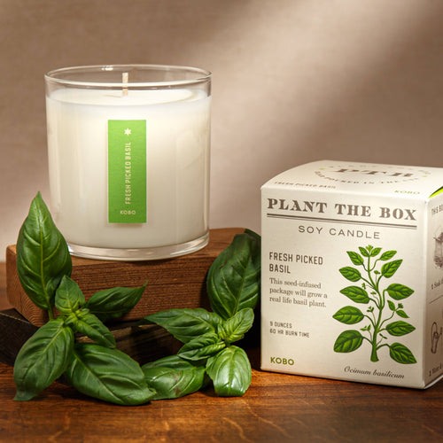 Close-up of a white soy candle with a lit wick, featuring a green basil label, on a wooden surface with a basil plant in the background.